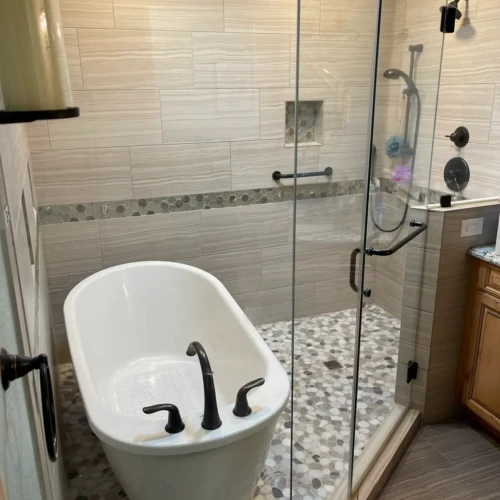 Luxury wet room in The Broadmoor, featuring a freestanding soaking tub next to a glass-enclosed walk-in shower with neutral wood-look tile and stone pebble floor by XCEL TILE.