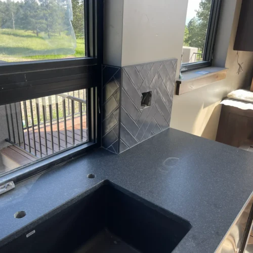 Kitchen window frame wrapped cleanly with dark grey herringbone pattern tile backsplash in Black Forest, CO, by XCEL TILE.