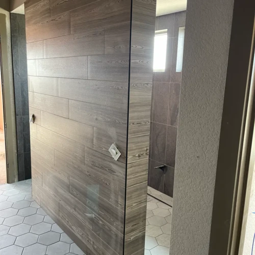 Custom bathroom partition wall in Castle Rock, CO, completely wrapped in horizontally stacked wood-look tile, contrasting with a light grey hexagon floor tile by XCEL TILE.