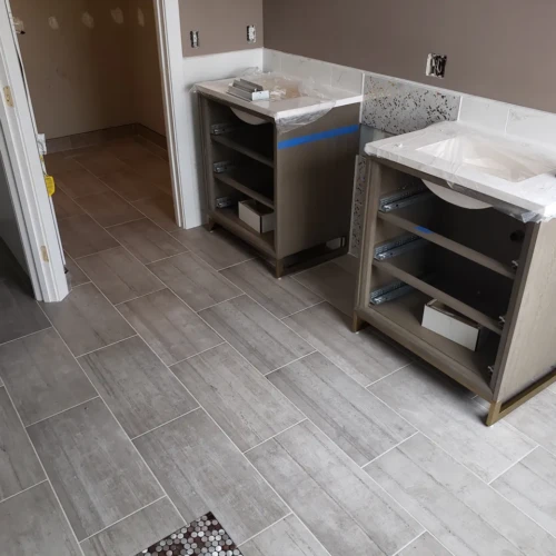 Residential bathroom under construction in Parker, CO, featuring two wood vanities installed over a continuous light grey wood-look tile floor that extends into a water closet by XCEL TILE.