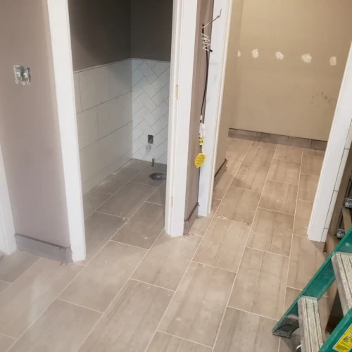 Wide view of a master bathroom remodel in Parker, CO, showing light grey wood-look tile flooring flowing seamlessly from the vanity area into a separate toilet room featuring a white herringbone wainscoting by XCEL TILE.